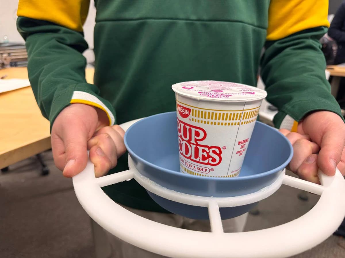 Two hands lifting the CoolTray prototype by its white ring handles, a blue bowl with a cup of instant noodles seated in the center.