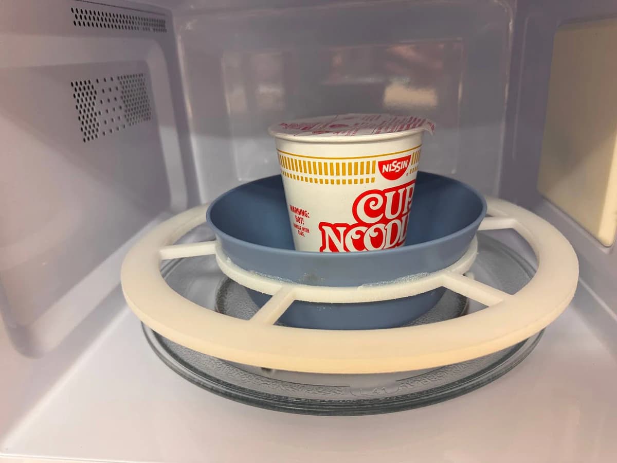 CoolTray prototype inside a microwave: a white ring frame with spokes holds a blue bowl, with a cup of instant noodles in the bowl.