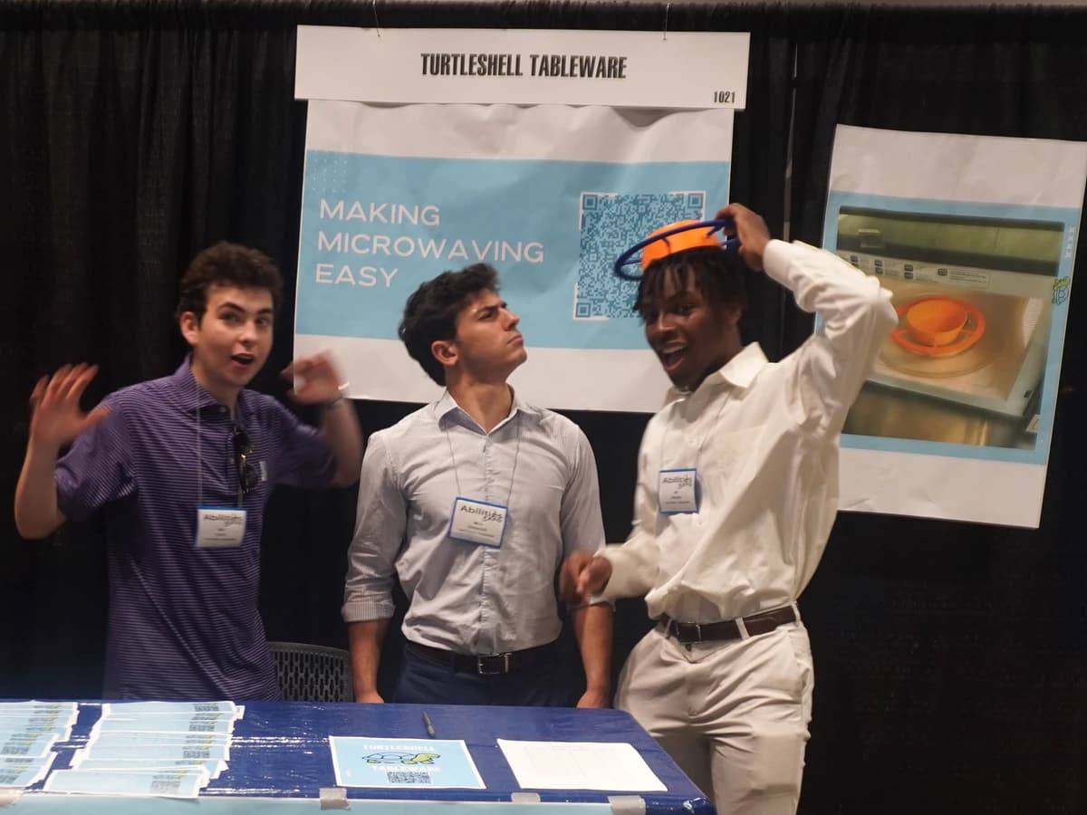 The three TurtleShell founders goofing around at their trade show booth under a 'Making microwaving easy' banner, one wearing a tray prototype as a hat, flyers on the table in front.
